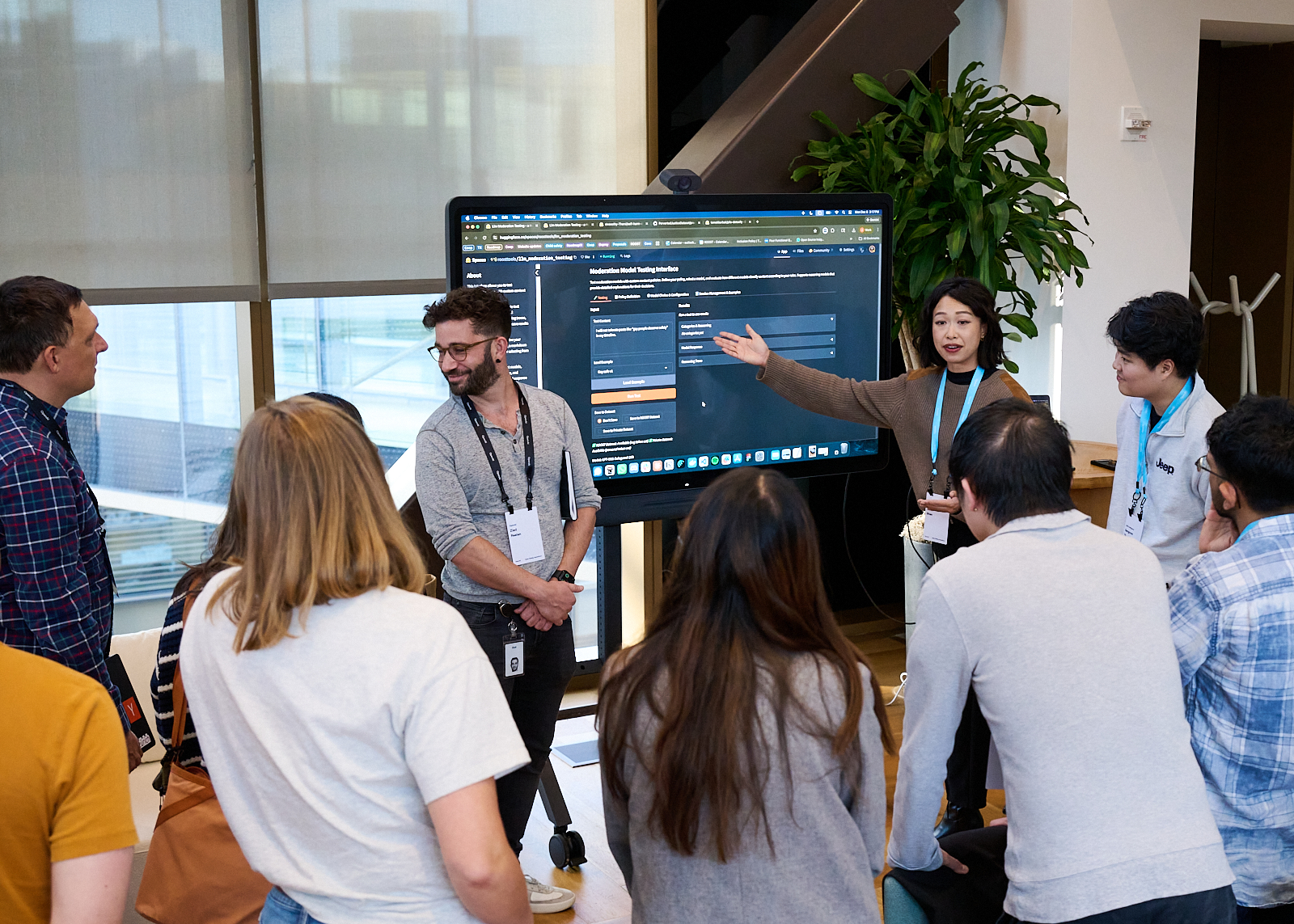 A group of people crowd around a large monitor looking at the screen showing an interface on HuggingFace for exploring the safety models. A woman in a brown sweater gestures towards the screen, explaining how it works.